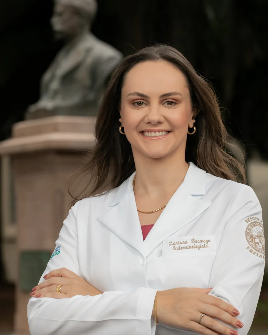 Dra. Larissa Basmage de braços cruzados, com jaleco branco bordado “Larissa Basmage — Endocrinologista”